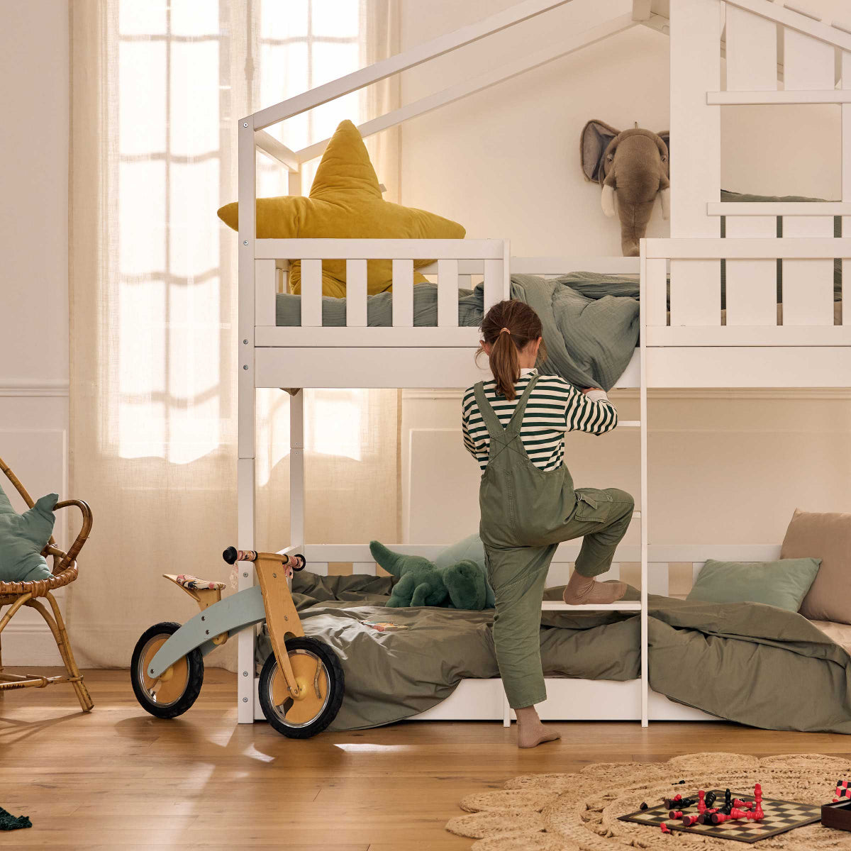 Chambre d'enfant chaleureuse avec un lit superposé cabane en bois blanc. Une fillette grimpe sur l'échelle pour rejoindre la couchette supérieure, décorée de coussins verts et jaunes. L'ambiance est douce et naturelle, avec un sol en bois, des rideaux beige et des éléments de décoration en matières naturelles. Une draisienne en bois et un tapis rond en jute complètent l'univers inspiré de la pédagogie Montessori.