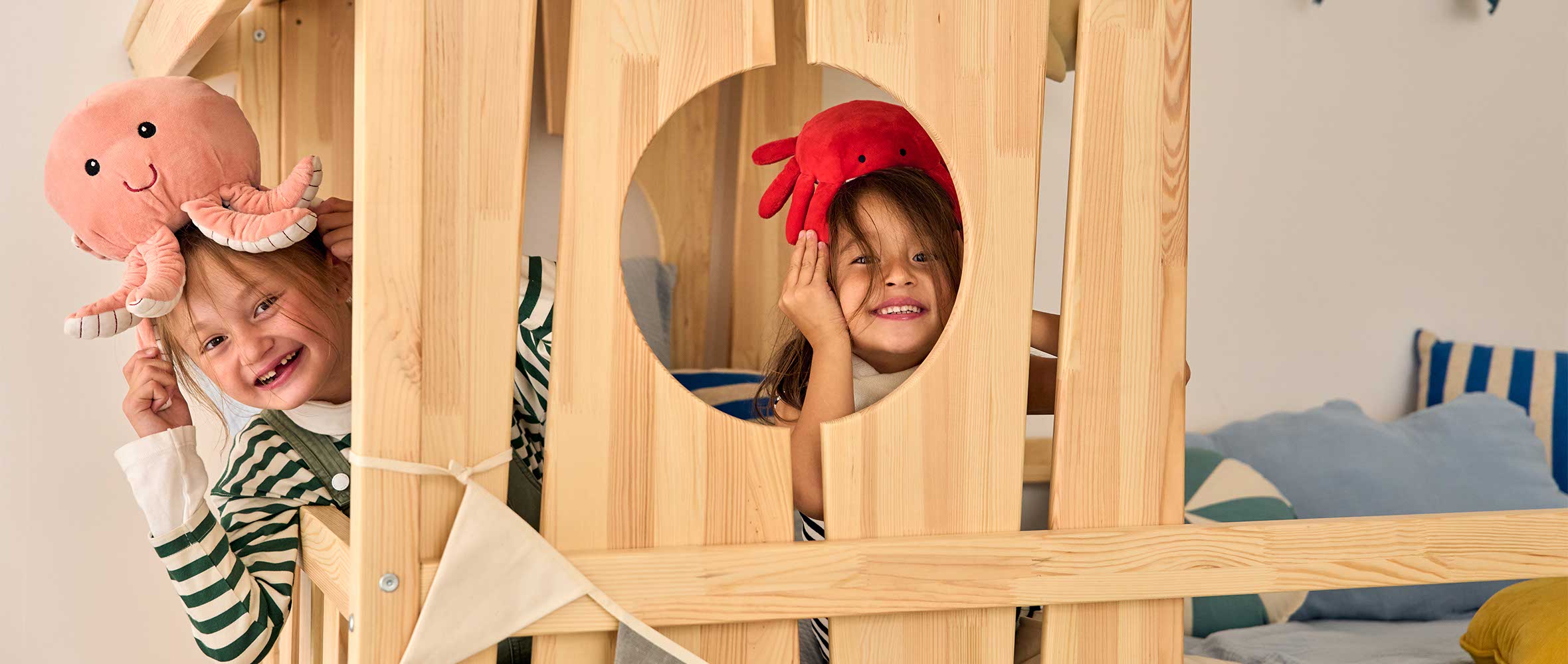 Deux enfants jouent dans un lit cabane en bois naturel, transformé en véritable refuge d'aventure. L'un porte une peluche pieuvre rose sur la tête, l'autre un crabe rouge, ajoutant une touche ludique et marine à l’ambiance. Le design du lit, avec sa fenêtre ronde et ses détails en bois brut, stimule l'imaginaire et invite aux jeux. En arrière-plan, des coussins aux tons bleus et rayés renforcent une atmosphère chaleureuse et réconfortante, idéale pour une chambre d’enfant inspirée par la nature et l’évasion
