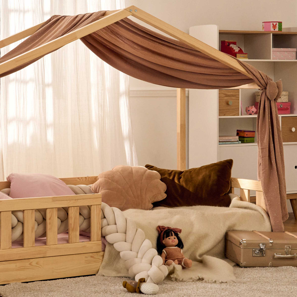 Chambre d'enfant cosy avec un lit cabane en bois naturel, agrémenté d'un voile en tissu brun noué sur le côté. L'espace est décoré avec des coussins en velours aux tons doux, une couverture moelleuse et une barrière tressée en textile. Une poupée en tissu et une valisette rétro apportent une touche chaleureuse. L'ambiance apaisante et bohème est sublimée par une lumière naturelle filtrant à travers des rideaux légers.